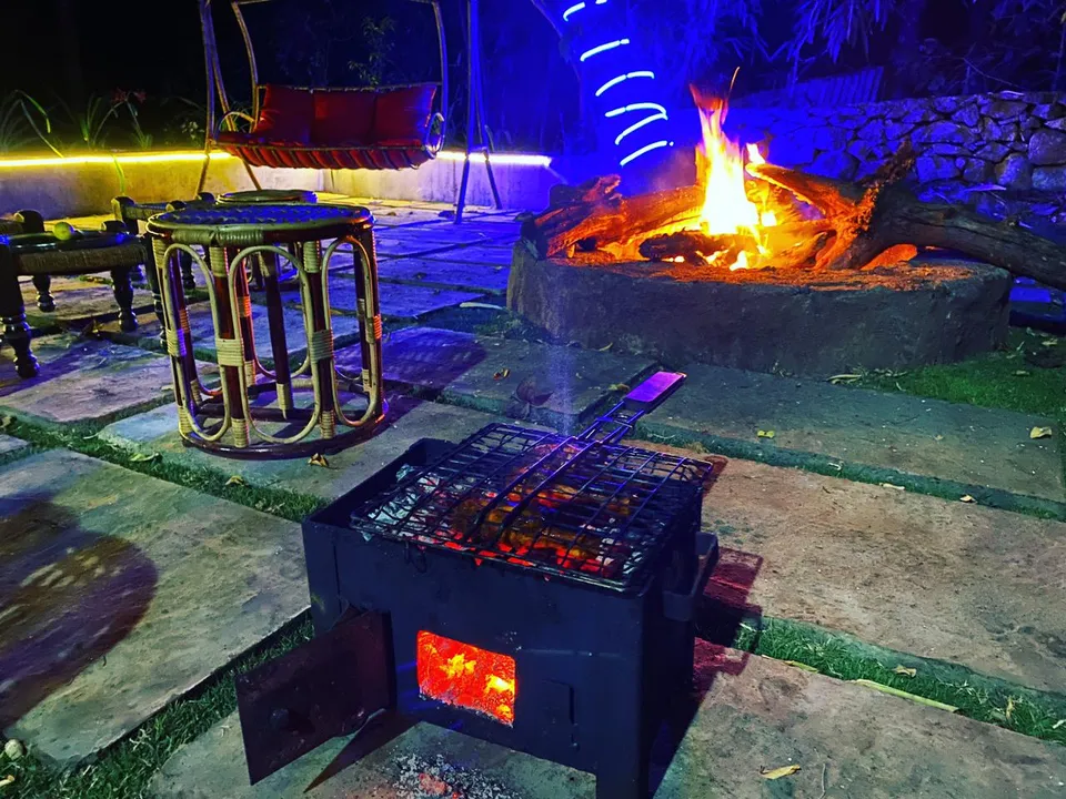 A log fire in a stone fire pit at night, with a small charcoal grill, cane stools and a cushioned swing