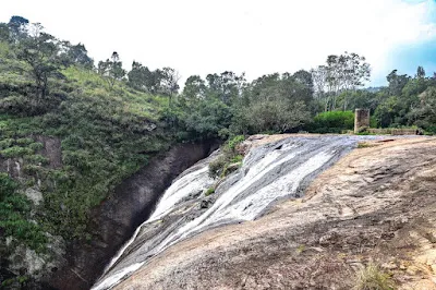 Water sheeting down a broad slope of rock into a gorge, with trees beyond